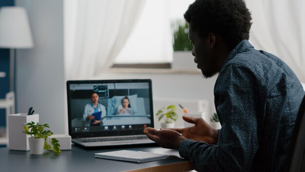 Messa a fuoco selettiva sul papà afroamericano preoccupato per la bambina nel reparto ospedaliero, parlando con il medico in videoconferenza con lo schermo remoto dell'app webcam per la connessione familiare. Consulente medico di famiglia