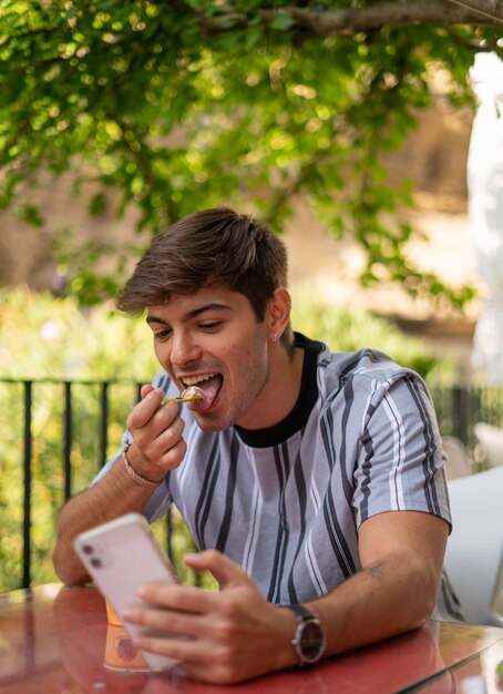 Messa a fuoco selettiva verticale di un bel giovane che mangia il gelato in un bar usando il suo telefono