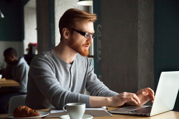 Messo a fuoco uomo millenario della testarossa che per mezzo del computer portatile che si siede al tavolo del caffè