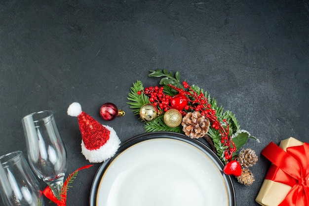 Metà colpo di piatto di cena albero di Natale rami di abete conifera cono confezione regalo cappello di Babbo Natale calici di vetro caduti su sfondo nero