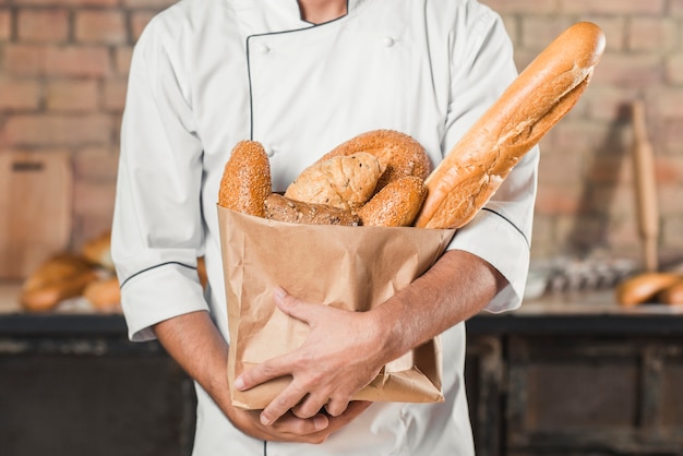 Metà di sezione del panettiere maschio che tiene diversi tipi di pane nel sacco di carta