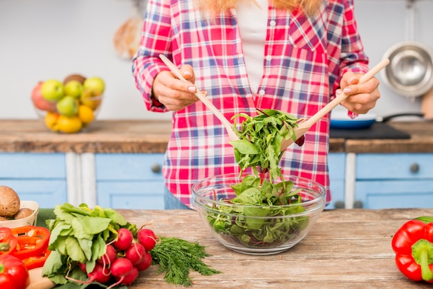 Metà di sezione di una donna che tiene insalata di verdure frondosa con il cucchiaio di legno sulla tavola di legno