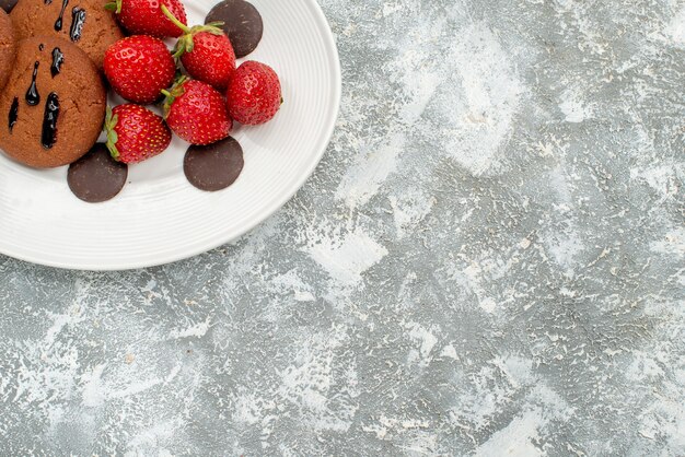 Metà superiore vista biscotti al cioccolato fragole e cioccolatini rotondi sulla piastra ovale bianca in alto a sinistra del fondo grigio-bianco