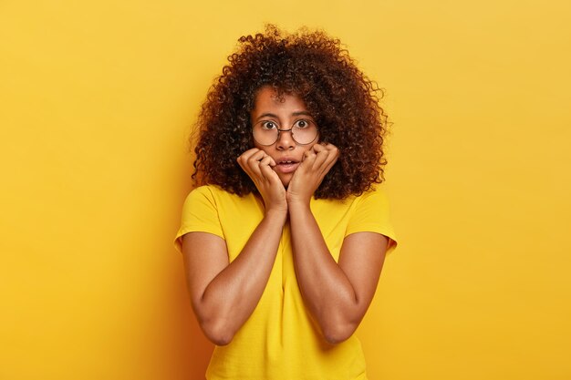 Mezza lunghezza colpo di ragazza imbarazzata con acconciatura afro, guarda con paura alla telecamera, tiene il mento, indossa occhiali da vista e maglietta gialla brillante, posa al coperto