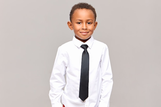 Mezzo busto bello allievo elementare maschio dalla pelle scura in posa isolato vestito con camicia bianca stirata e cravatta elegante nera, pronto per andare a scuola, sorridente