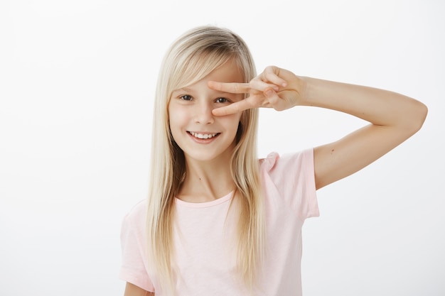 Mezzo busto di affascinante bambino positivo con i capelli biondi in abbigliamento casual, che mostra la vittoria o il gesto di pace sugli occhi e sorride felice, balla o si diverte sul muro grigio, è di ottimo umore