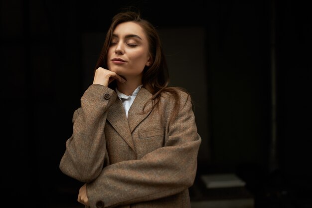 Mezzo busto di attraente giovane donna europea alla moda con lunghi capelli castani in posa isolato tenendo gli occhi chiusi, con espressione facciale pacifica sognante, indossando giacca da uomo, sorridente