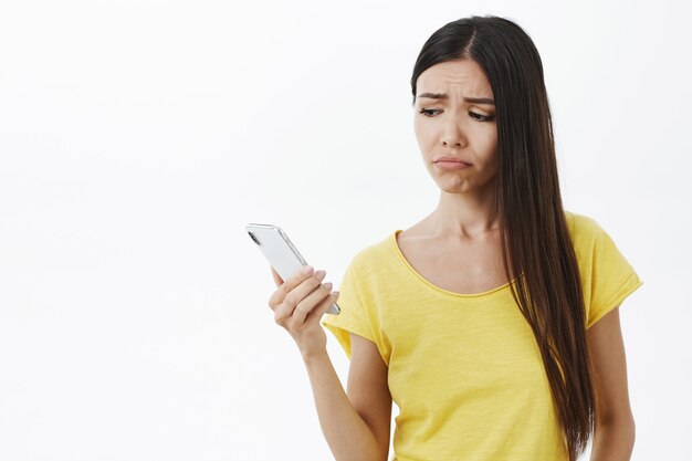 Mezzo busto di bella giovane donna sconvolta infelice senza trucco in maglietta gialla che tiene smartphone guardando lo schermo del cellulare