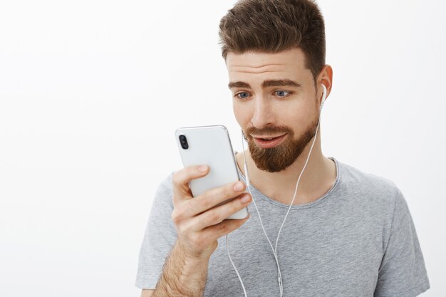 Mezzo busto di carino affascinante maschio barbuto con gli occhi azzurri che indossa gli auricolari tenendo lo smartphone vicino al viso durante la lettura o la visione di toccare il video affascinante guardando il cellulare felice