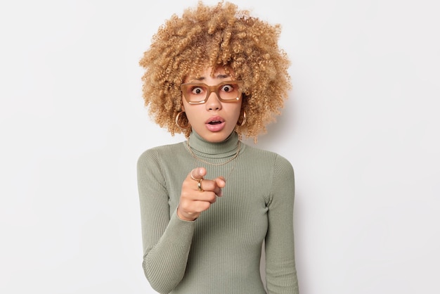 Mezzo busto di donna bionda dai capelli ricci scioccata trattiene il respiro dalla meraviglia non può credere che i suoi occhi indichino in avanti con il dito indice indossa dolcevita casual isolato su sfondo bianco