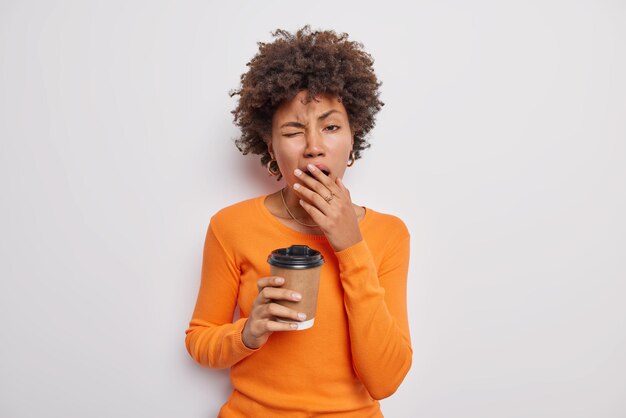 Mezzo busto di donna bruna riccia assonnata stanca copre la bocca e sbadigli beve caffeina bevanda per rinfrescare indossa un maglione arancione casual isolato su sfondo bianco. Persone e concetto di stanchezza