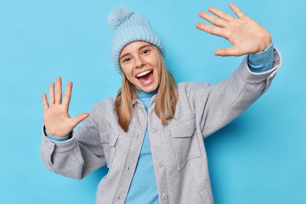 Mezzo busto di felice donna spensierata tiene i palmi alzati verso la telecamera inclina la testa esclama con gioia indossa un caldo cappello invernale la giacca grigia si diverte isolato su sfondo blu sembra pieno di gioia