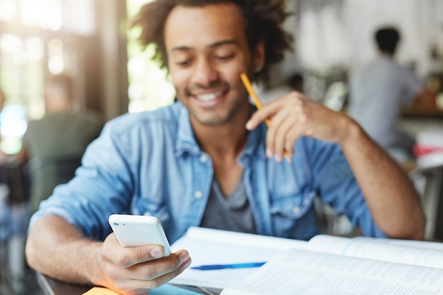 Mezzo busto di felice studente di college afroamericano con sorriso carino digitando un messaggio di testo su gadget elettronico, seduto al tavolino del bar con libri di testo. Messa a fuoco selettiva sulla mano dell'uomo che tiene il telefono cellulare