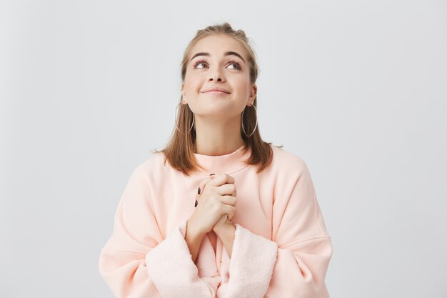 Mezzo busto ritratto di femmina caucasica allegra con capelli biondi in posa in studio con lo sguardo verso l'alto. Ragazza vestita in felpa rosa che tiene i palmi premuti insieme, con l'accattonaggio.