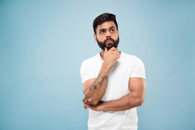 Mezzo busto vicino ritratto di giovane uomo indù in camicia bianca su sfondo blu. Emozioni umane, espressione facciale, concetto di annuncio. Spazio negativo. Pensando tenendo la mano sulla barba. Scegliere.