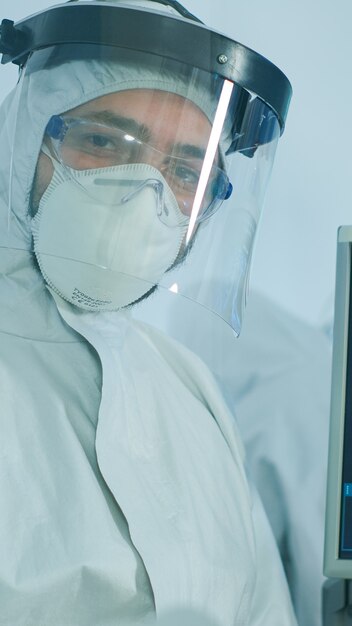 Microbiologo in tuta dpi in piedi in laboratorio guardando la telecamera dietro la parete di vetro nel laboratorio attrezzato. Medico che esamina l'evoluzione del virus utilizzando strumenti ad alta tecnologia e chimica per lo sviluppo di vaccini
