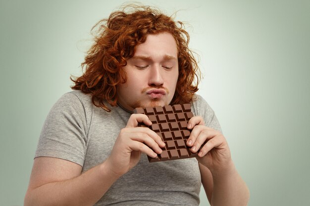 Mio prezioso! Ritratto di divertente giovane dai capelli rossi in sovrappeso dipendente dallo zucchero che indossa una maglietta grigia con una grande barra di cioccolato con entrambe le mani, imbronciato le labbra, non vedo l'ora di mordere un po '