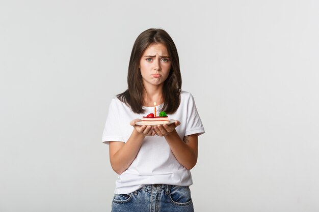 Miserabile ragazza di compleanno che sembra triste, tenendo la torta di compleanno con la candela.