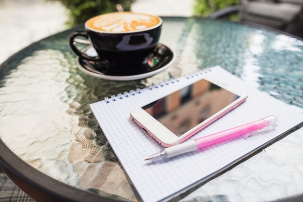 Mobile sul notebook controllato e penna con caffè su un tavolo all&#39;aperto