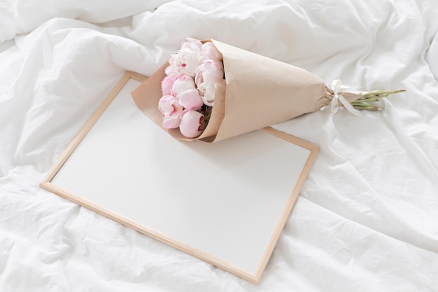 Mockup con cornice bianca sul letto Bouquet di peonie rosa in confezione artigianale Interni bianchi scandinavi