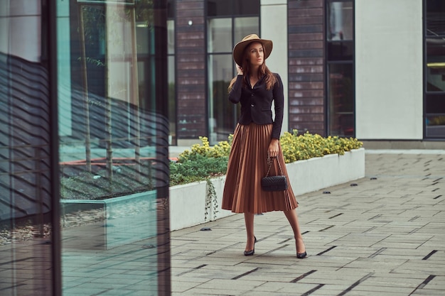 Moda donna elegante che indossa una giacca nera, cappello marrone e gonna con una pochette a mano che cammina su un centro cittadino europeo.