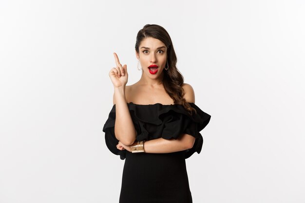 Moda e bellezza. Attraente donna caucasica in abito nero che ha un'idea, alzando il dito e dicendo suggerimento, sfondo bianco.