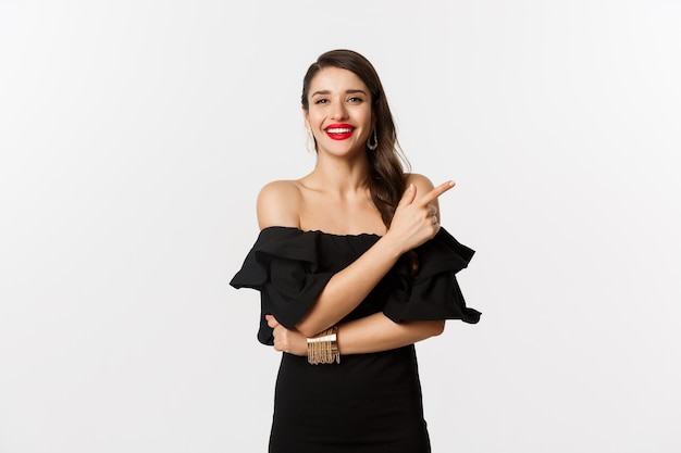 Moda e bellezza. Donna elegante con labbra rosse, vestito nero, sorridente e puntando il dito proprio sul logo, in piedi su sfondo bianco.