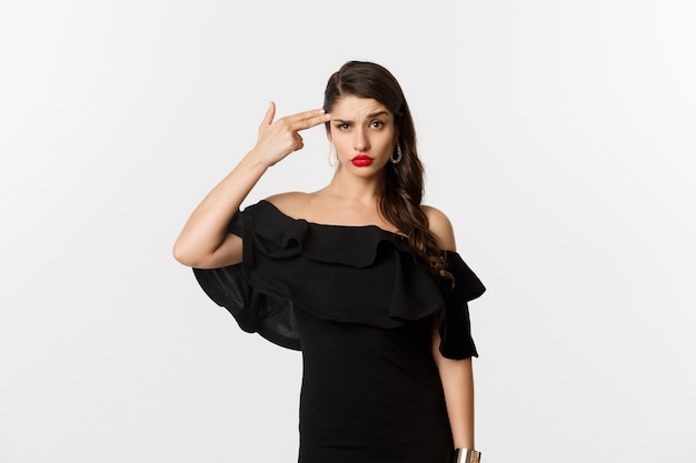 Moda e bellezza. Giovane donna infastidita in vestito nero che fa il gesto della pistola del dito vicino alla testa, sparandosi dall'irritazione, stando sopra il fondo bianco.