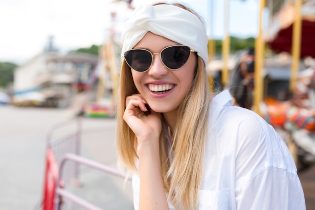 Moda giovane donna dolce sorridente con capelli biondi che ride alla macchina fotografica che indossa una camicia bianca e accessori per capelli bianchi e occhiali da sole neri sulla strada da attrazioni