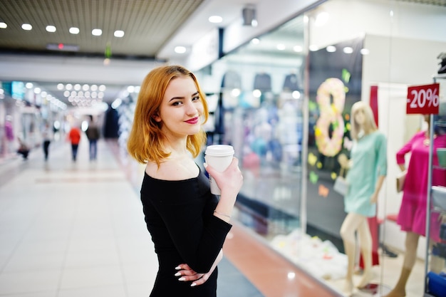Moda ragazza dai capelli rossi indossa un abito nero con una tazza di caffè luminosa che tiene in mano una tazza di caffè al centro commerciale commerciale Filtri Instagram in stile foto tonica