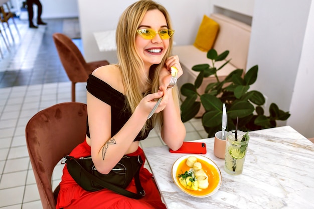 Moda ritratto al coperto di giovane donna bionda alla moda hipster in posa al caffè moderno hipster, colazione del mattino.