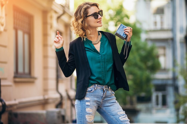 Moda ritratto di giovane donna elegante che cammina per strada in giacca nera, camicetta verde, accessori eleganti, borsetta in mano, occhiali da sole, stile fashion street estivo