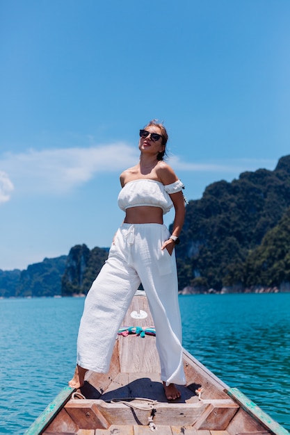 Moda ritratto di giovane donna in top bianco e pantaloni in vacanza, sulla barca a vela in legno tailandese. Concetto di viaggio. Femmina nel parco nazionale di Khao Sok.