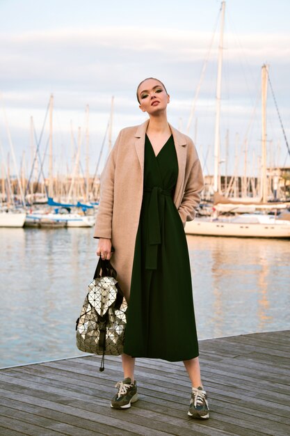 Moda ritratto di giovane splendida donna elegante in posa lungomare, indossando scarpe da ginnastica cappotto abito e zaino, turista di lusso, morbidi colori caldi.