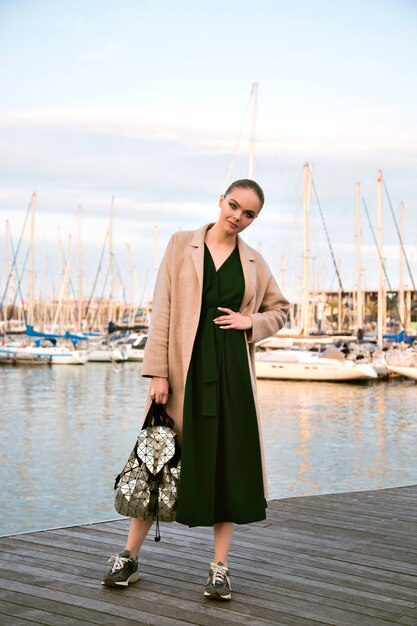 Moda ritratto di giovane splendida donna elegante in posa lungomare, indossando scarpe da ginnastica cappotto abito e zaino, turista di lusso, morbidi colori caldi.