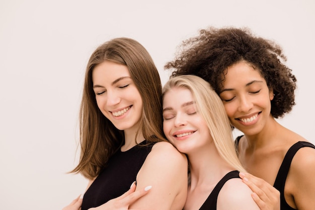 Modelli multietnici di tre giovani donne con gli occhi chiusi in cime nere in piedi insieme su sfondo bianco