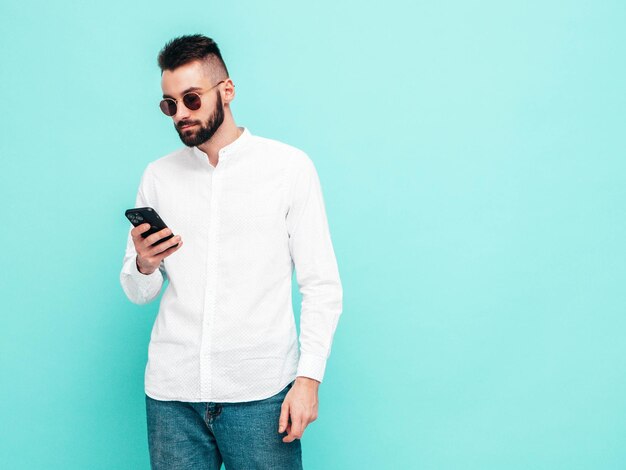 Modello bello e sicuro di sé Uomo sexy ed elegante vestito con camicia e jeans Moda hipster maschio in posa vicino al muro blu in studio Holding smartphone Guardando lo schermo del cellulare Usando app isolate