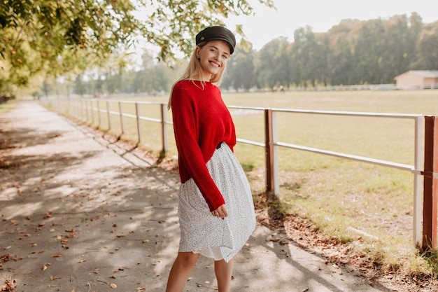 Modello biondo felice che sembra trendy in vestiti casuali rossi. Bella donna che mostra un sorriso allegro sulla foto nel parco.