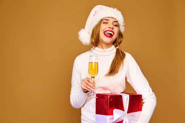 Modello con cappello da Babbo Natale e confezione regalo grande bevendo champagne sul muro dorato