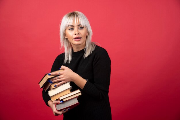 Modello della giovane donna che tiene molti libri su una parete rossa.