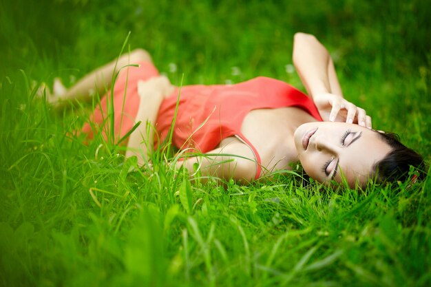 modello di bella donna castana che si trova nell'erba luminosa di estate verde nel parco con trucco in vestito rosso.