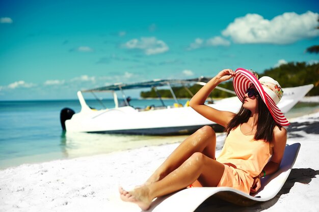 Modello di bella donna che prende il sole sulla sedia di spiaggia in bikini bianco in cappellino da sole variopinto dietro l'oceano blu dell'acqua di estate