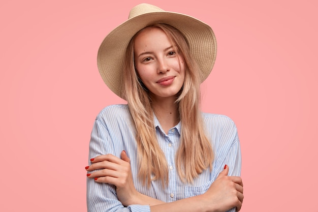 Modello di bella e timida giovane donna felice che tiene le mani incrociate, guarda positivamente la telecamera, indossa cappello e camicia