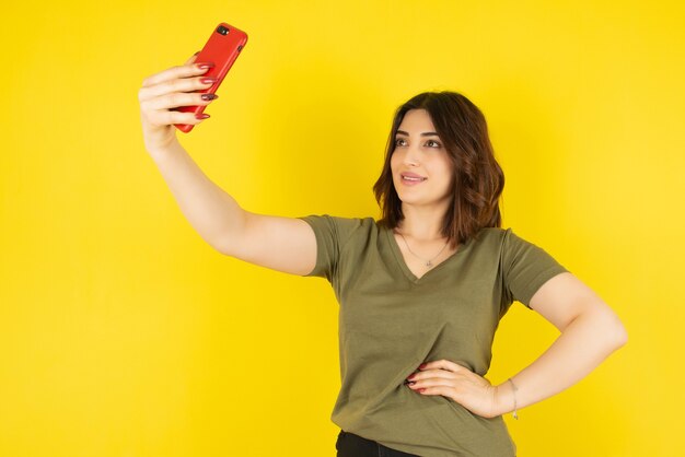 Modello di donna castana in piedi e prendendo selfie con il suo telefono cellulare contro il muro giallo