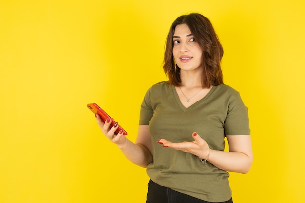 Modello di donna castana in piedi e usando il suo telefono cellulare contro il muro giallo.