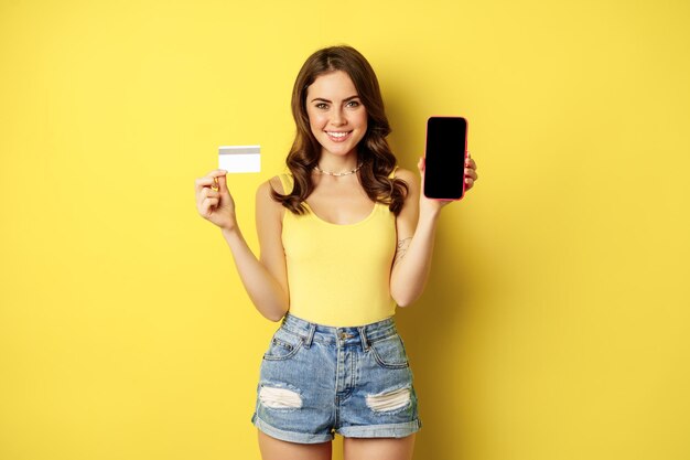Modello di giovane bella donna che mostra lo schermo del telefono vuoto dello smartphone e la carta di credito, pronto per l'estate, indossando canotta e pantaloncini, in piedi su sfondo giallo.