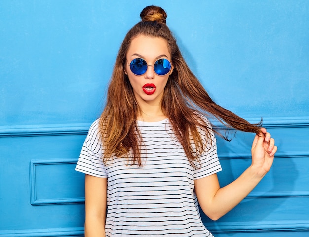 Modello di giovane donna alla moda in abiti casual estivi con labbra rosse, in posa vicino alla parete blu. Giocando con i suoi capelli