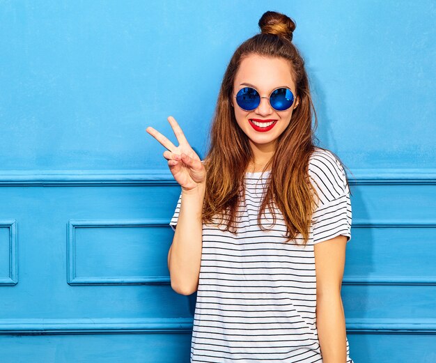 Modello di giovane donna alla moda in abiti casual estivi con labbra rosse, in posa vicino alla parete blu. Mostra del segno di pace