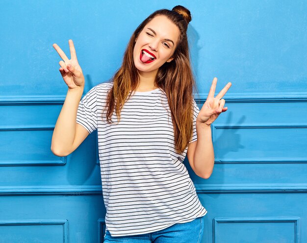 Modello di giovane donna alla moda in abiti casual estivi con labbra rosse, in posa vicino alla parete blu. Strizza l'occhio e mostra il segno di pace