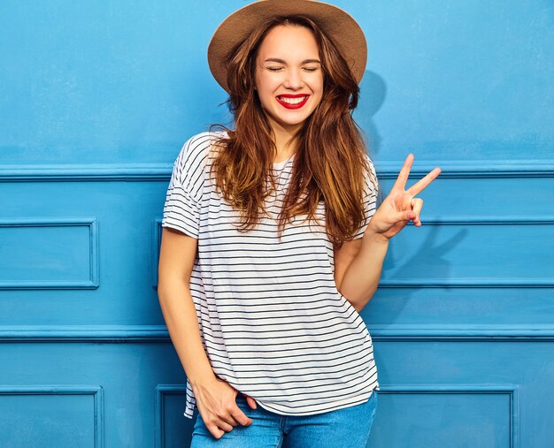 Modello di giovane donna alla moda in abiti casual estivi e cappello marrone con labbra rosse, in posa vicino alla parete blu. Mostra del segno di pace e della sua lingua